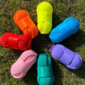 Puede incluir: Siete porta-hilos de colores en forma de arcoíris. Los porta-hilos son de plástico y tienen una superficie texturizada. Cada porta-hilos tiene una pequeña etiqueta con un gráfico.
