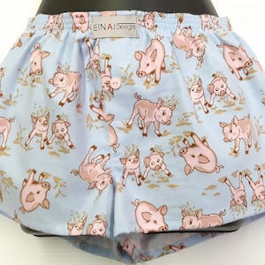 May include: Light blue boxer shorts with a pattern of pink piglets. The shorts have a button fly and an elastic waistband. The label on the shorts reads "EINAI Designs".