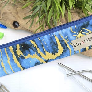 May include: A blue and gold zippered pouch with the words 'EINAI Designs' printed on it. The pouch is made of fabric with a marble pattern and is lying on a wooden surface.
