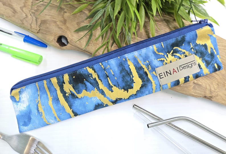 May include: A blue and gold zippered pouch with the words 'EINAI Designs' printed on it. The pouch is made of fabric with a marble pattern and is lying on a wooden surface.