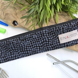 May include: A black and gray zippered pouch with a white label that reads 'EINAI Designs'. The pouch has a black and gray polka dot pattern. The pouch is lying on a white surface with a fork and two metal straws.