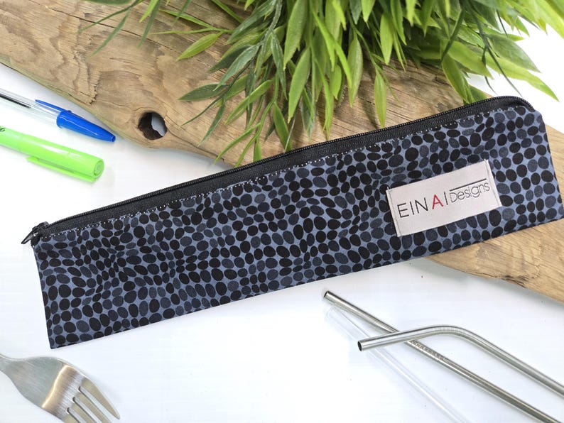 May include: A black and gray zippered pouch with a white label that reads 'EINAI Designs'. The pouch has a black and gray polka dot pattern. The pouch is lying on a white surface with a fork and two metal straws.