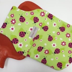 May include: A red rubber hot water bottle with a green and white fabric cover. The cover has a pattern of ladybirds and flowers. The text "EINAI Designs" is printed on the cover.