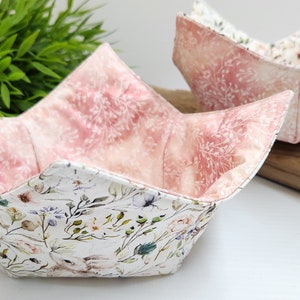 May include: Two fabric bowl cozies in a pink and white floral pattern. The cozies are folded and displayed on a wooden surface.