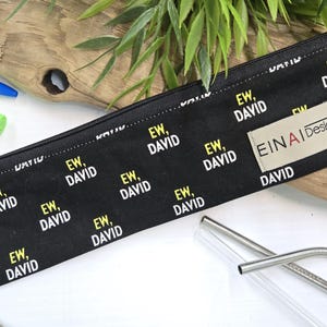 May include: A black zippered pouch with a repeating pattern of the text 'EW, DAVID' in white and yellow. The pouch has a beige label with the text 'EINAI Designs' in black. The pouch is lying on a white surface with two clear glass drinking straws.