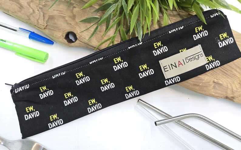 May include: A black zippered pouch with a repeating pattern of the text 'EW, DAVID' in white and yellow. The pouch has a beige label with the text 'EINAI Designs' in black. The pouch is lying on a white surface with two clear glass drinking straws.