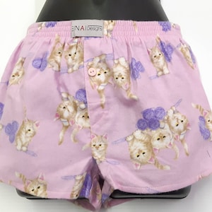 May include: Pink cotton pyjama shorts with a button fly fastening and a repeating pattern of kittens holding balls of yarn. The shorts have the text "EINAI Designs" printed on the waistband.
