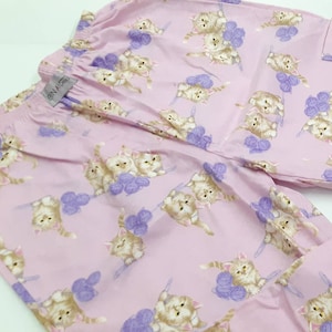 May include: Pink pyjama trousers with a pattern of kittens playing with balls of yarn. The trousers have a waistband with an elastic band and a drawstring. The label on the trousers reads "Ein-A-Way".