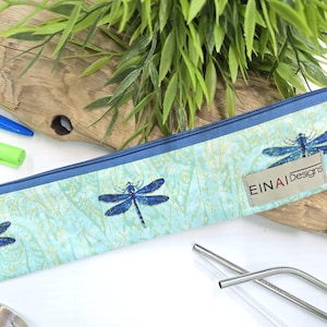 May include: A blue zippered pouch with a dragonfly pattern and the text 'EINAI Designs' on it. The pouch is lying on a wooden surface with two metal straws and a clear glass straw next to it.