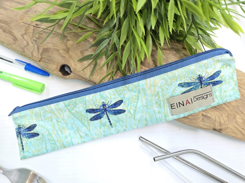 May include: A blue zippered pouch with a dragonfly pattern and the text 'EINAI Designs' on it. The pouch is lying on a wooden surface with two metal straws and a clear glass straw next to it.