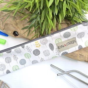 May include: A white fabric pouch with a zipper closure and a gray trim. The pouch has a repeating pattern of green, gray, and yellow stylized trees. The pouch has a label that reads 'EINAI Designs'. Two stainless steel drinking straws are visible in the foreground.