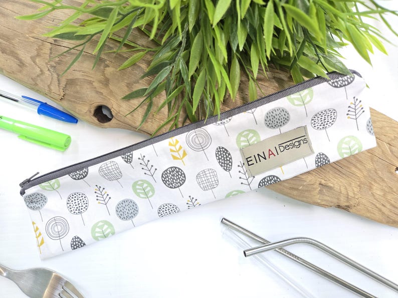 May include: A white fabric pouch with a zipper closure and a gray trim. The pouch has a repeating pattern of green, gray, and yellow stylized trees. The pouch has a label that reads 'EINAI Designs'. Two stainless steel drinking straws are visible in the foreground.