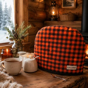 May include: A dome-shaped tea cosy featuring a vibrant red and black buffalo check pattern. The fabric cover includes a small white rectangular label with the black text EINAI Designs stitched onto the lower right corner of the exterior surface.