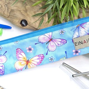 May include: A blue zippered pouch with a butterfly print and the text "EINAI Designs". The pouch is made of fabric and has a zipper closure. The pouch is lying on a wooden surface.