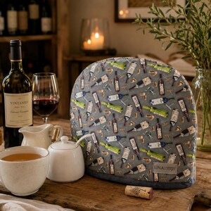 May include: A grey fabric tea cosy featuring a repeating pattern of wine bottles, corkscrews, and corks. The cosy has a rounded shape with a dark grey base trim and a small rectangular label with the text EINA Design stitched on the lower right side.