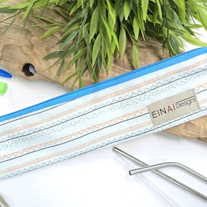 May include: A blue and white striped fabric pouch with a zipper closure. The pouch has a small tan label with the text 'EINAI Designs'. The pouch is lying on a wooden surface with two stainless steel drinking straws next to it.