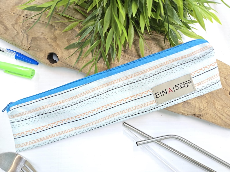 May include: A blue and white striped fabric pouch with a zipper closure. The pouch has a small tan label with the text 'EINAI Designs'. The pouch is lying on a wooden surface with two stainless steel drinking straws next to it.