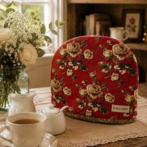 May include: A dome-shaped tea cosy featuring a deep red fabric base with a repeating pattern of cream-coloured roses and green leaves. A small white rectangular tag with the black text EINA Designs is sewn onto the lower right corner of the cosy.