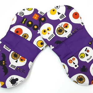 Peut inclure: Une paire de maniques de cuisine violettes pour Halloween avec un fond blanc et un motif de crâne et d'os croisés. Les maniques portent les mots "Boo Boo" et "eek" imprimés dessus.