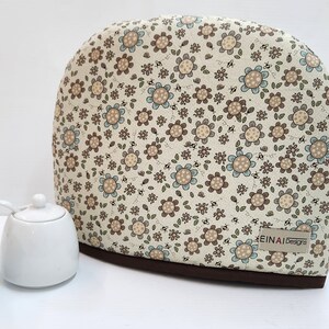 May include: A beige teapot cosy with a floral pattern of brown and blue flowers. The cosy has a brown trim and the text "EINAI Designs" is printed on the bottom right corner.