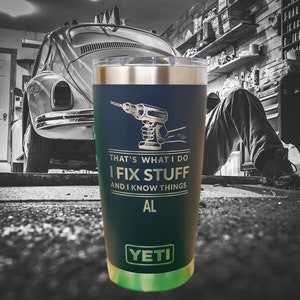 Peut inclure: Un gobelet YETI bleu foncé avec une inscription dorée qui dit "That's what I do I fix stuff and I know things AL". Le gobelet présente une image dorée d'une perceuse et se trouve devant une voiture ancienne dans un garage.