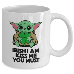 Puede incluir: Taza de cer&aacute;mica blanca con un personaje de dibujos animados verde que sostiene un tr&eacute;bol de cuatro hojas. El texto "IRISH I AM KISS ME YOU MUST" est&aacute; impreso en la taza.