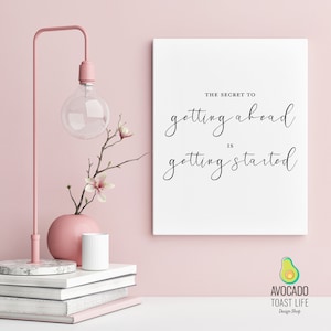 May include: A white canvas print with black calligraphy that reads "The secret to getting ahead is getting started." The print is on a pink wall with a pink lamp, a vase with white flowers, and a stack of books.