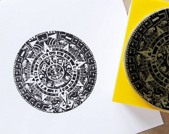 May include: A black and white rubber stamp featuring a detailed design of the Aztec calendar, also known as the Sun Stone. The stamp is mounted on a yellow block.