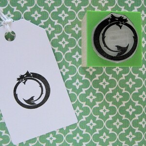 Op de afbeelding: Een wit label met een zwart Ouroboros-ontwerp, een draak die zijn eigen staart opeet, hangt aan een touwtje. Een bijpassende stempel ligt op een groen blok. De achtergrond is een groen en wit patroon.