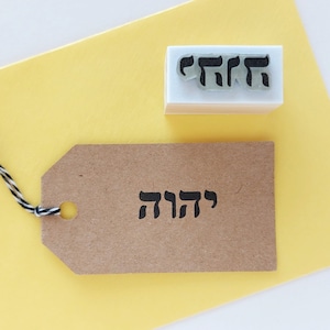 Pode incluir: Uma etiqueta de papel castanho com a palavra hebraica "שלום" (que significa "paz") carimbada nela. Também é mostrado um carimbo de borracha branco com a mesma palavra hebraica.