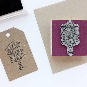 May include: A rubber stamp featuring a Tree of Life design with Hebrew text, mounted on a square wooden block with a purple foam base. Next to it is a brown paper tag stamped with the same black ink Tree of Life design and a black ink pad.
