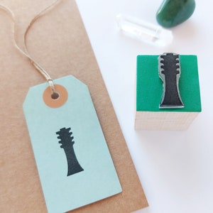 Op de afbeelding: Een houten blok met een groene bovenkant en een zwart silhouet van een gitaar erop. Het blok staat op een wit oppervlak naast een bruine papieren zak met een blauw label eraan. Het label heeft ook een zwart silhouet van een gitaar erop.