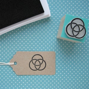 Könnte beinhalten: Ein blauer Holzblock mit einem schwarzen Stempel aus Gummi mit drei ineinandergreifenden Kreisen. Ein braunes Papieretikett mit einem schwarzen Stempel aus Gummi mit drei ineinandergreifenden Kreisen. Ein weißes Stempelkissen mit einem schwarzen Stempelkissen.