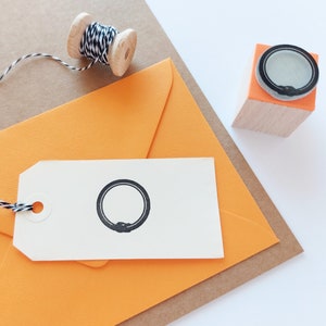 May include: A white tag with a black circle design is tied to an orange envelope with twine. A wooden stamp with a black circle design is on a brown surface.