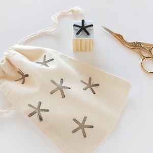 Op de afbeelding: Een witte katoenen trekkoordtas met een zwart sterrenpatroon. De tas ligt naast een houten blokstempel met een zwart sterrenpatroon en een gouden schaar.