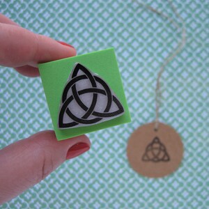 May include: A green rubber stamp with a black and white Celtic knot design. The stamp is held in a hand.