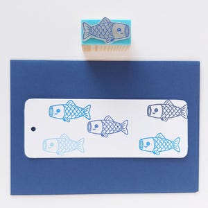 May include: A blue fish stamp with a wooden handle sits above a navy blue card with a white tag. The tag features multiple blue fish illustrations. The stamp and illustrations are in a cartoon style.