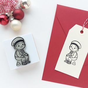 May include: A square rubber stamp featuring a line art illustration of a child wearing a cap and vest, sitting next to a small pile. A matching stamped cream-colored paper tag is attached to a red envelope with red and white baker's twine.