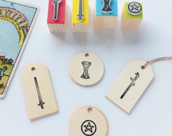 May include: Four wooden stamps with red, yellow, blue, and green tops featuring wand, sword, chalice, and pentacle symbols. Below are matching wooden tags and circular discs stamped with the same black ink designs, alongside a tarot card.