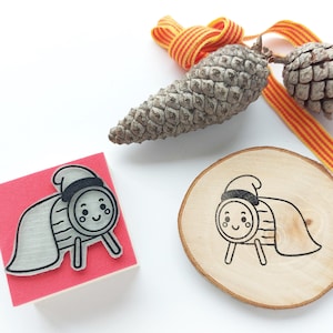 以下が含まれることがあります： 赤いスタンプと木の円盤には、帽子とマントを着た漫画のキャラクターが描かれています。松ぼっくりとオレンジと黄色のストライプのリボンも画像にあります。キャラクターは笑顔です。