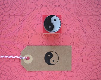 May include: A brown paper gift tag with a black and white yin yang symbol and a red and white string attached to it. The tag is sitting on a pink and white patterned background.