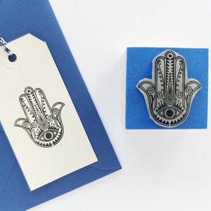 May include: A blue rubber stamp featuring a detailed black Hamsa hand design with an eye at the center, positioned next to a cream-colored paper gift tag with the same Hamsa hand design stamped in black ink, attached to a blue envelope.