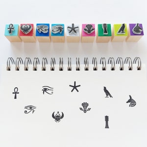 May include: A set of 10 rubber stamps featuring Egyptian symbols, including an ankh, eye of Horus, scarab beetle, and a star. The stamps are displayed on a white background in two rows. The stamps are colourful and have a wooden base.