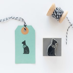 May include: A black and white rubber stamp with a design of a black cat wearing a collar. The stamp is on a gray square base. A teal tag with a black cat design is also shown. A spool of black and white twine is in the background.