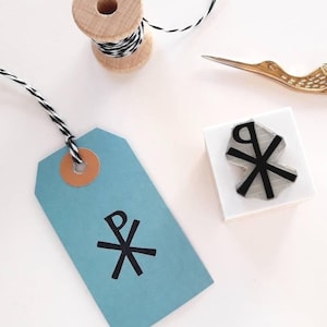 Peut inclure: Une étiquette bleu clair avec un symbole noir et un œillet marron, reliée à une bobine en bois de ficelle noire et blanche. Des ciseaux dorés et un tampon avec un symbole assorti sont également présents.