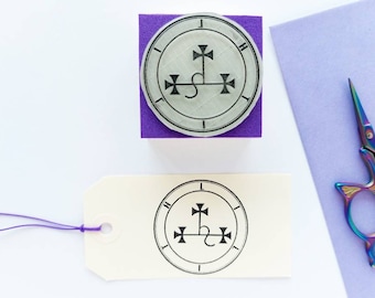 May include: A square purple rubber stamp featuring a circular design with a central cross symbol and the letters L, I, G, H, T arranged around the perimeter. Below the stamp is a cream-colored paper tag with the same black ink design stamped on it.