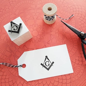 May include: A Masonic symbol stamp, a wooden spool of black and white twine, and vintage scissors rest on a patterned coral surface. A white tag with the same Masonic symbol is also present, suggesting a crafting or stationery theme.