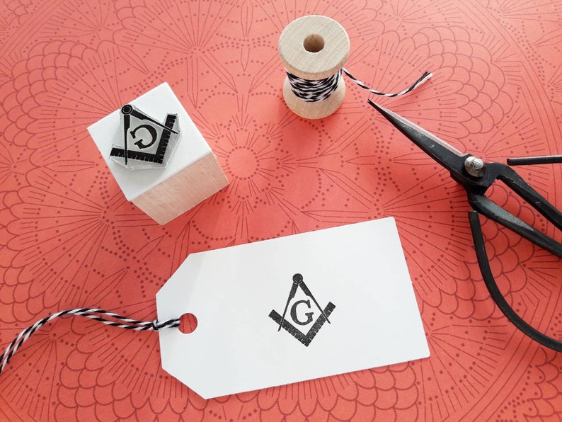 May include: A Masonic symbol stamp, a wooden spool of black and white twine, and vintage scissors rest on a patterned coral surface. A white tag with the same Masonic symbol is also present, suggesting a crafting or stationery theme.