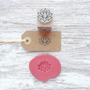 May include: A wooden rubber stamp featuring a lotus flower design, a brown paper gift tag with a black stamped lotus flower, and a circular piece of pink clay with an embossed lotus flower impression, all displaying the same floral motif.