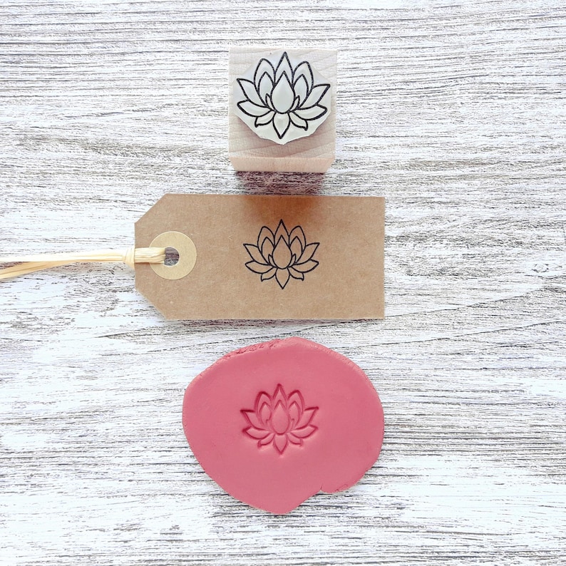 May include: A wooden rubber stamp featuring a lotus flower design, a brown paper gift tag with a black stamped lotus flower, and a circular piece of pink clay with an embossed lotus flower impression, all displaying the same floral motif.